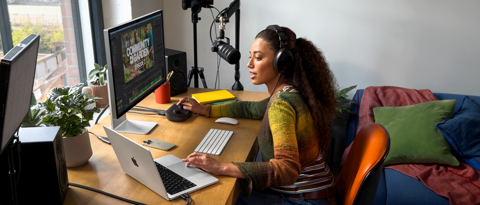 自宅のデスクで作業する女性。MacBook Proを2台の外部ディスプレイに接続し、ヘッドフォンを着けてマイクに向かって話している