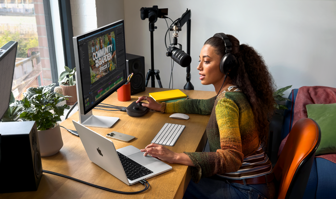 自宅のデスクで作業する女性。MacBook Proを2台の外部ディスプレイに接続し、ヘッドフォンを着けてマイクに向かって話している