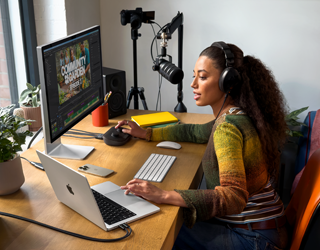 自宅のデスクで作業する女性。MacBook Proを2台の外部ディスプレイに接続し、ヘッドフォンを着けてマイクに向かって話している